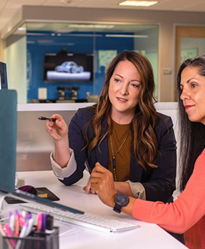 Two female colleagues collaborate at an office desk, with one pointing a pen toward a computer monitor.
