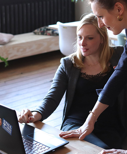 Two women collaborate at a wooden desk, focused on a laptop screen